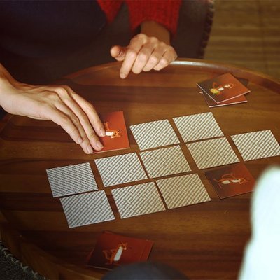 Auf dem Bild ist ein Memory Spiel auf einem brauen Holztisch mit einer Hand zu sehen, die eine Karte umdreht